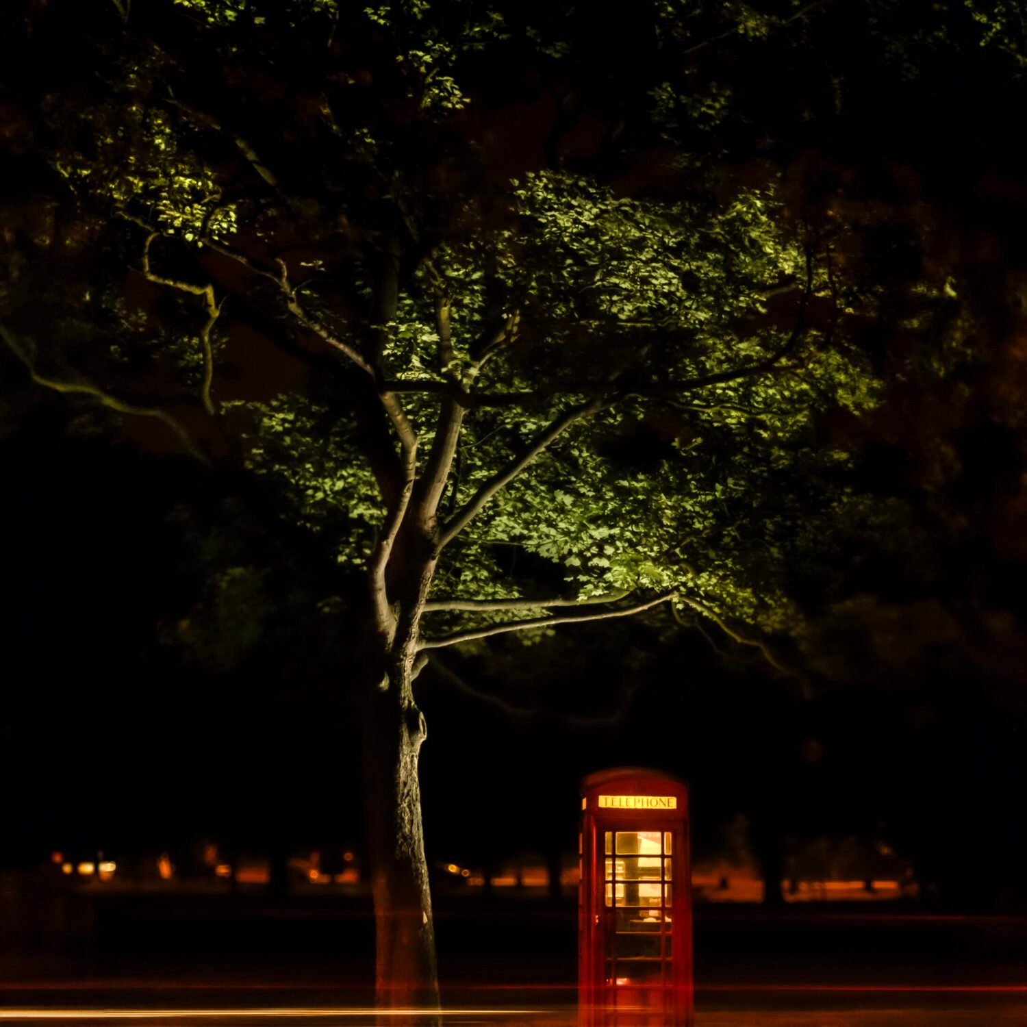 A traditional British red telephone box illuminated by night under a tree with passing traffic lights creating streaks in the foreground.