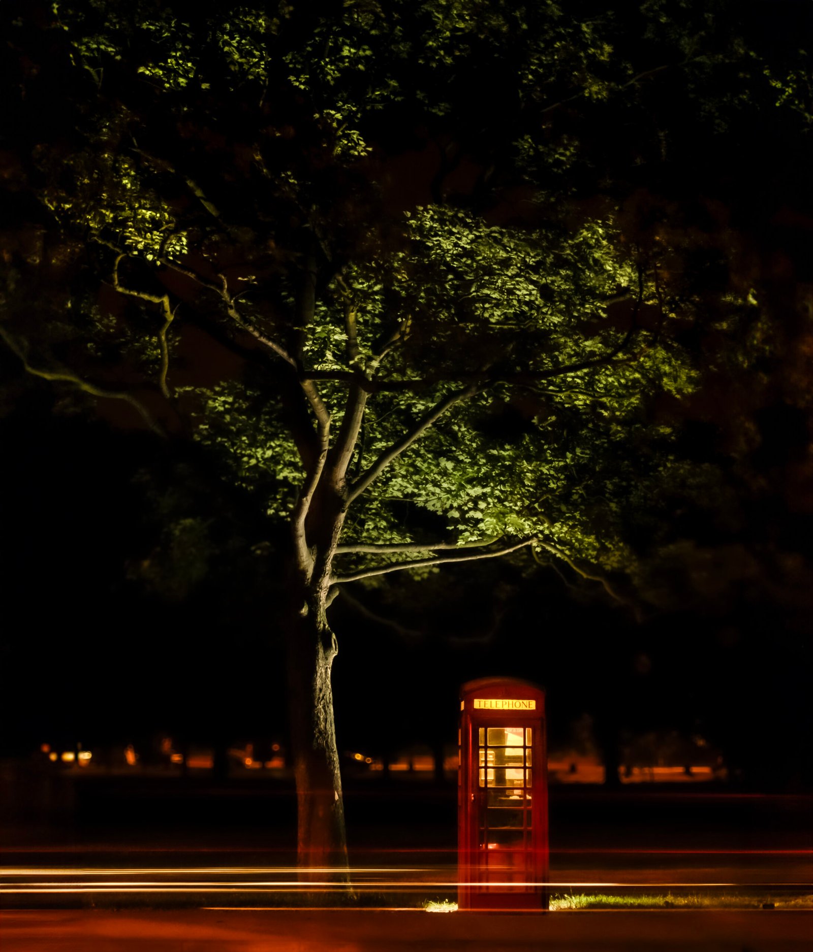 A traditional British red telephone box illuminated by night under a tree with passing traffic lights creating streaks in the foreground.