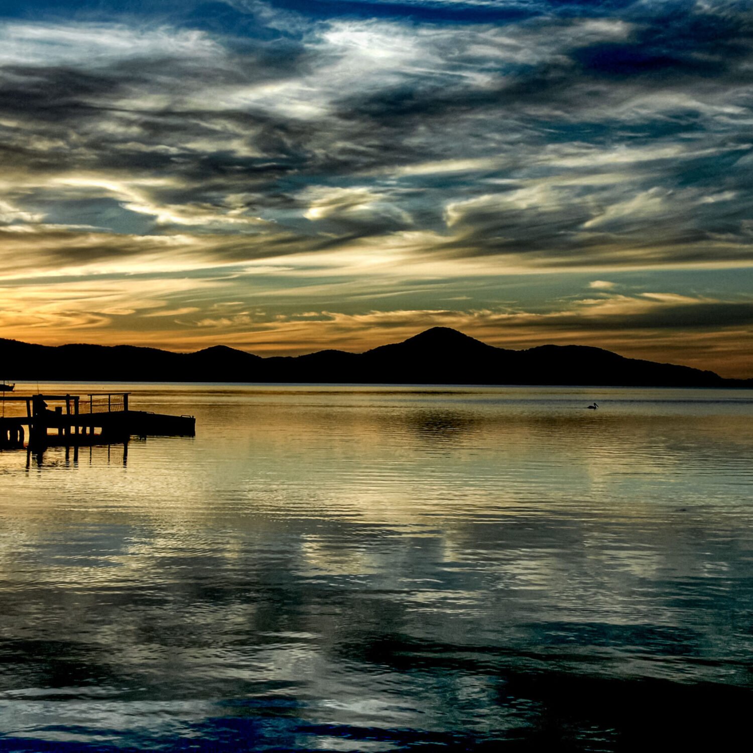 Golden sunset over a serene lake with silhouettes of a pier and distant mountains under a dramatic sky.