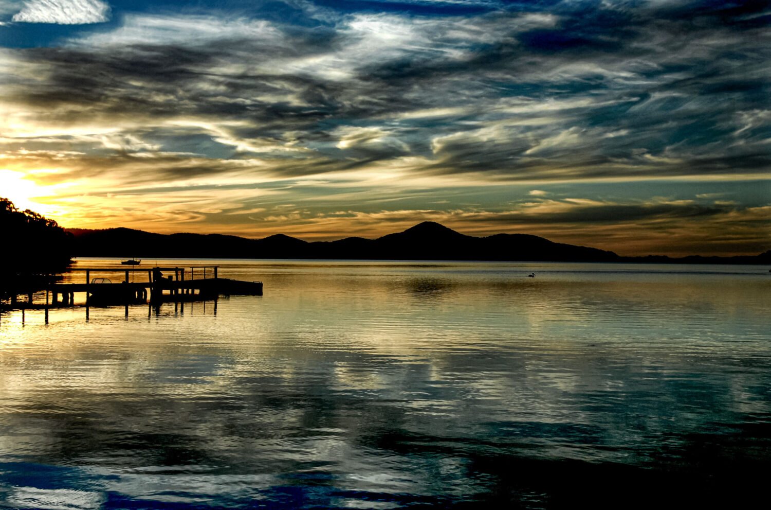 Golden sunset over a serene lake with silhouettes of a pier and distant mountains under a dramatic sky.