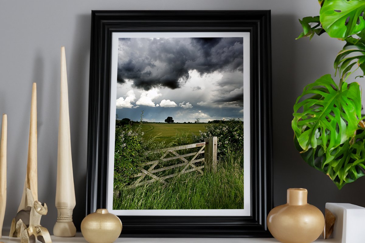 A wooden gate leading to a green field under a dramatic cloudy sky.